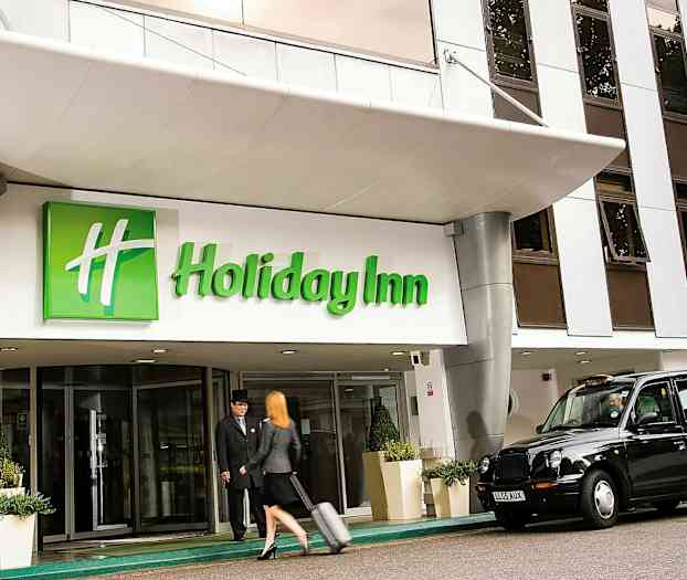 Inngangen til Holiday Inn med en taxi parkert utenfor og en person som drar en koffert. (AI-generert tekst)