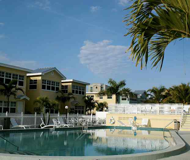 Et basseng omgitt av solsenger, parasoller og hus, med tropiske palmer i bakgrunnen. (AI-generert tekst)