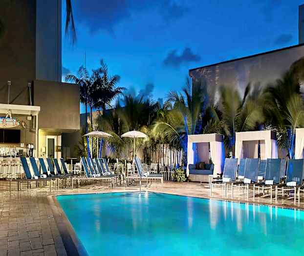 Et utendørs basseng med solsenger og cabanas rundt, samt en bar i bakgrunnen med palmer rundt. (AI-generert tekst)