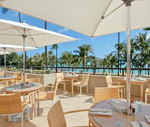 En uteterrasse med bord og stoler under parasoller, med havutsikt og palmer i bakgrunnen. (AI-generert tekst)