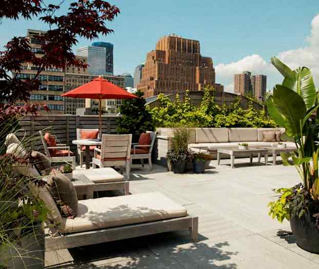 En romslig takterrasse med utemøbler, parasoll og planter. I bakgrunnen sees bygninger og en klar himmel. (AI-generert tekst)