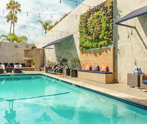 Et utendørs basseng med solsenger, grønne planter på veggen, og sitteplasser med puter. (AI-generert tekst)