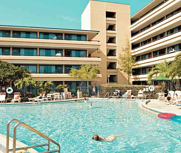 Et stort svømmebasseng omgitt av solsenger og en bygning med flere etasjer i bakgrunnen. (AI-generert tekst)