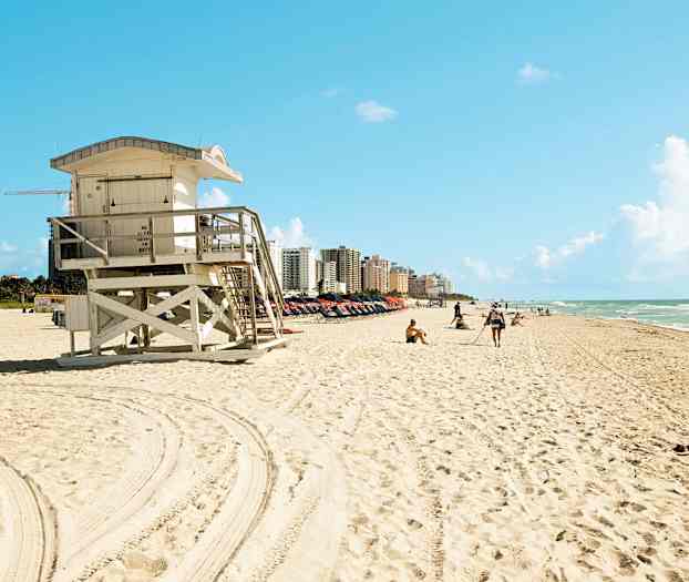 En strand med et livvaktstårn i forgrunnen og bygninger i bakgrunnen. (AI-generert tekst)