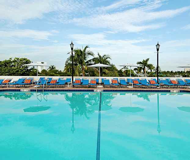 Et stort utendørs basseng med solsenger og parasoller langs kanten, omgitt av palmer. (AI-generert tekst)