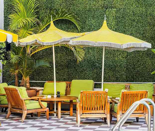 Utendørsstoler med grønne puter og trebord under gul- og hvitstripede parasoller, med vegetasjon i bakgrunnen. (AI-generert tekst)