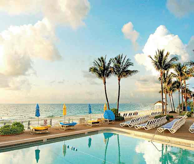 Et basseng med solsenger og parasoller ved sjøen, med palmer i bakgrunnen. (AI-generert tekst)