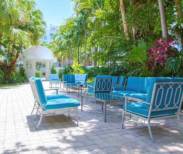 Utemøbler med blå puter står plassert på en steinbelagt uteplass, omgitt av palmer og grønn vegetasjon. (AI-generert tekst)