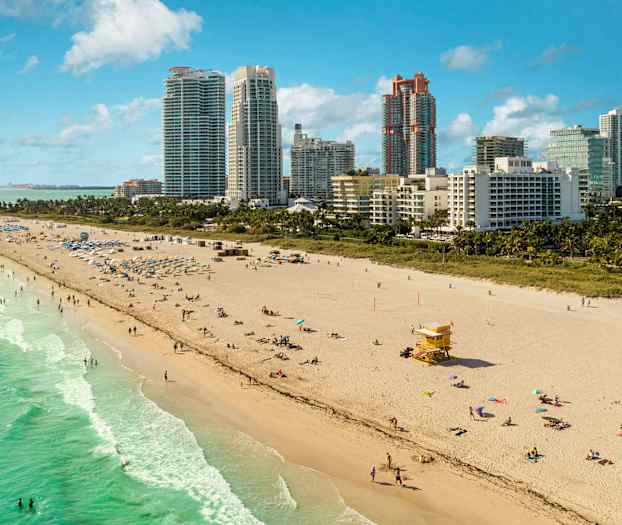 En sandstrand med noen mennesker som bader og soler seg nær sjøen. Høye bygninger er synlige i bakgrunnen. (AI-generert tekst)