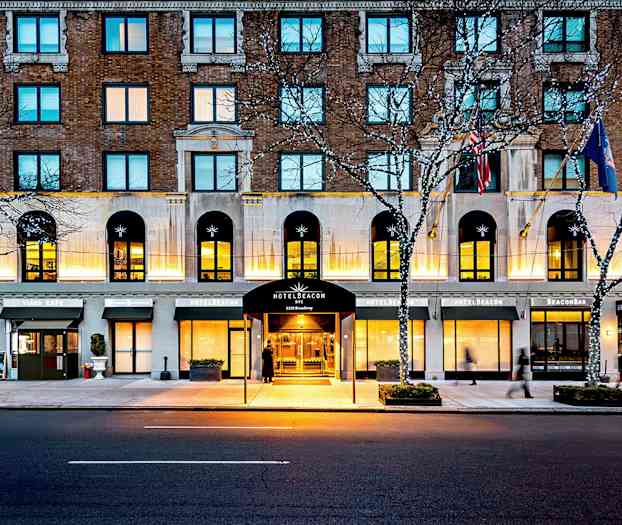 Hotellbygning ved en gate, med inngang merket som Hotel Beacon, omgitt av kafémiljø og lyslenker i trær. (AI-generert tekst)