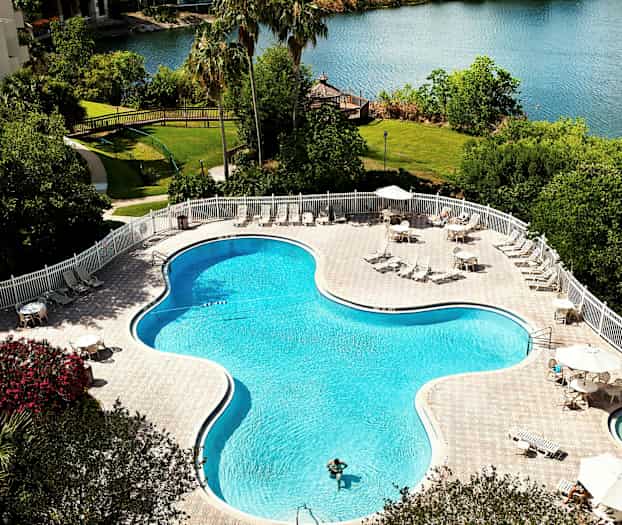 Et svømmebasseng utendørs omgitt av solsenger og bord med parasoller ved en innsjø. (AI-generert tekst)