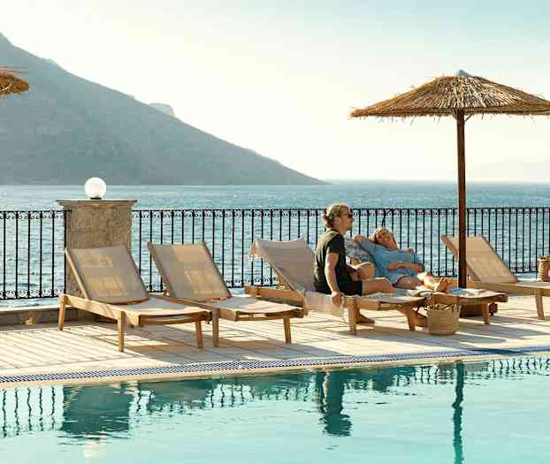 Solstoler ved et basseng med utsikt over havet og fjell i bakgrunnen. (AI-generert tekst)