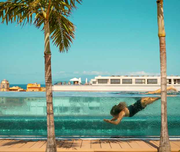 En person svømmer under vann i et gjennomsiktig basseng, med bygninger og en palme mot en blå himmel i bakgrunnen. (AI-generert tekst)