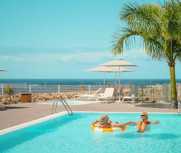 Et basseng med solsenger og parasoller ved siden av, mot en bakgrunn av hav og palmer. (AI-generert tekst)