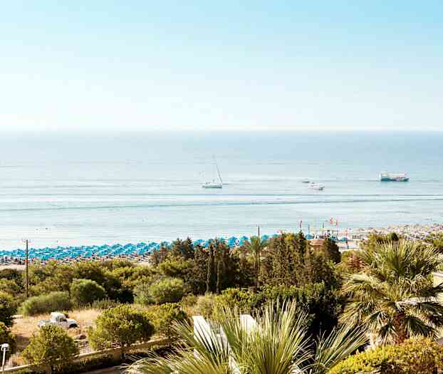 Utsikt over en strand med blå parasoller, omgitt av grønne trær. Flere båter kan sees på det rolige havet. (AI-generert tekst)