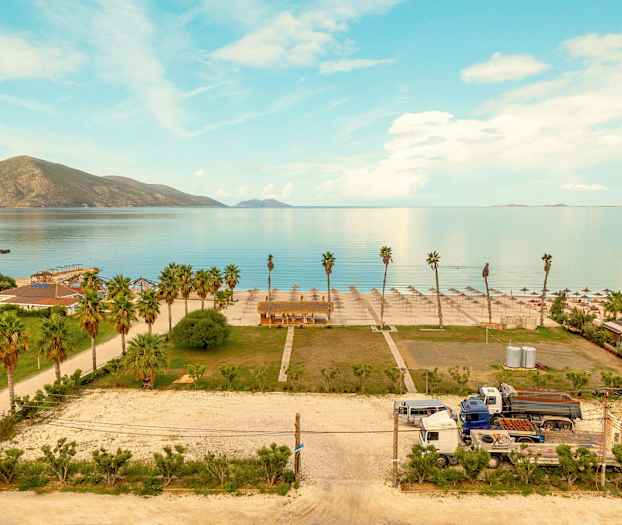 Utsikt over en strand med parasoller og palmer, en del av bygninger og kjøretøy er også synlige. (AI-generert tekst)