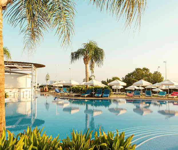 Et basseng omgitt av solsenger med blå og røde puter, parasoller og palmer. (AI-generert tekst)