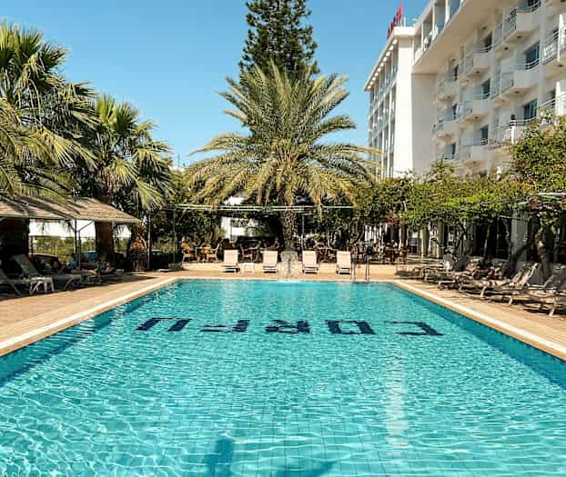 Utendørs svømmebasseng med solsenger og palmer, ved siden av en hotellbygning. (AI-generert tekst)