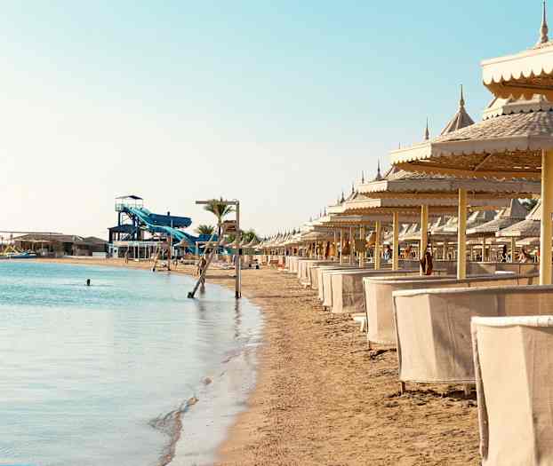 En sandstrand med solsenger og parasoller, samt vannsklier i bakgrunnen. (AI-generert tekst)