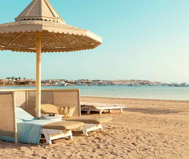 En strand med solsenger og parasoller, samt båter på havet. (AI-generert tekst)