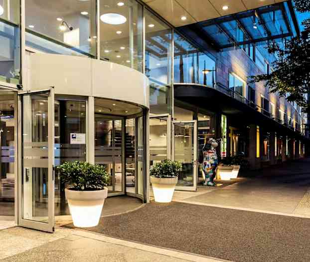 Inngangen til en bygning med glassdører og roterende dør, omgitt av store blomsterkrukker og opplyst under kveldens belysning. (AI-generert tekst)