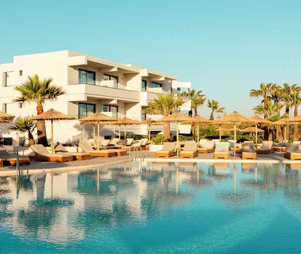 Hotell med hvit fasade bak et svømmebasseng og solsenger med parasoller. (AI-generert tekst)