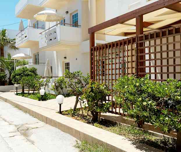 En gate med hvite bygninger, grønne busker og parasoller. (AI-generert tekst)