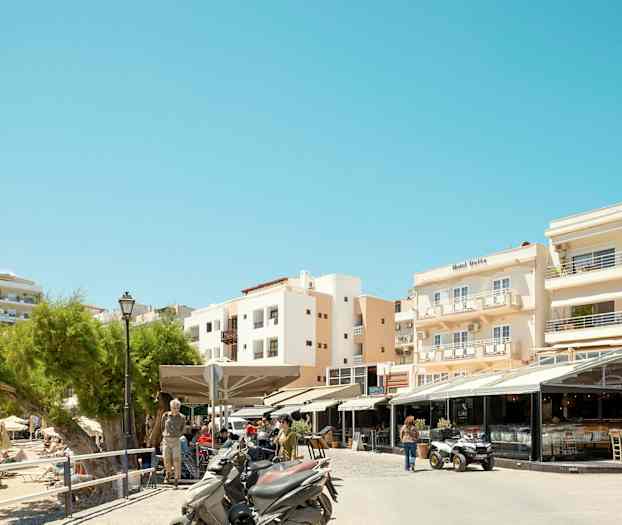 Bilde av en gate med restauranter og motorsykler foran bygninger i et solrikt område. (AI-generert tekst)