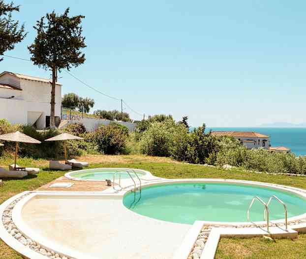 Et utendørsbasseng med solsenger og parasoller, omgitt av gress og vegetasjon. Sjøen og bygninger er synlige i bakgrunnen. (AI-generert tekst)