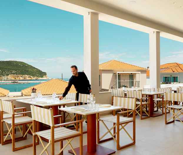 Restaurant med bordoppsett, utsikt over havet og terrasse, med bakgrunn av terrakottatak og en frodig åsside. (AI-generert tekst)