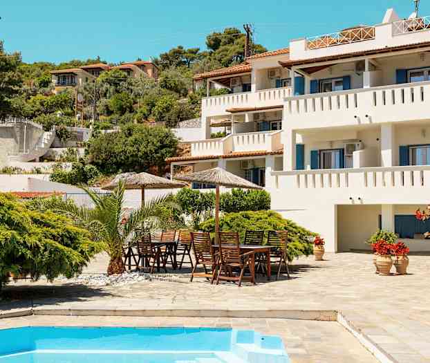 Hvit bygning med balkonger, frodig hage og basseng i forgrunnen. (AI-generert tekst)