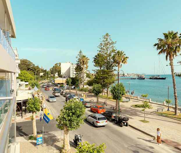 En bygateg som går ved siden av havet, med biler, palmer og noen bygninger i bakgrunnen. (AI-generert tekst)