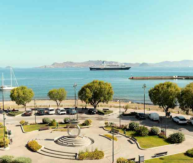 Utsikt over en havn med parkeringsplass og trær i forgrunnen, båter og et cruiseskip på det blå havet, samt fjell i bakgrunnen. (AI-generert tekst)