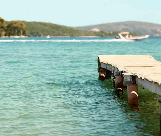 En trebrygge som strekker seg ut i klart vann, med en motorbåt og frodige åser i bakgrunnen. (AI-generert tekst)