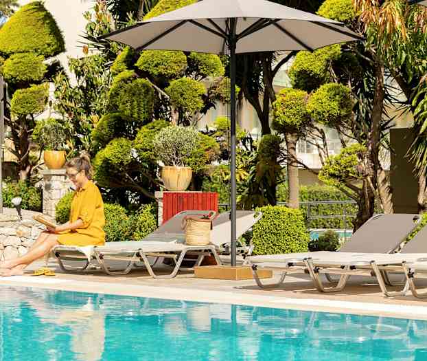 En kvinne i gul kjole sitter med en åpen bok ved et basseng, omgitt av solsenger og parasoller. (AI-generert tekst)