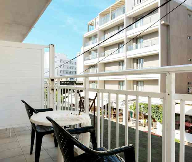 En balkong med to stoler og et bord, utsikt mot bygninger. (AI-generert tekst)