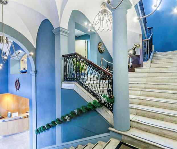 Et interiørbilde av en hall med en trapp i lys marmor og blå vegger, dekorert med grønne girlandere. Trappen har rekkverk av svart metall og trematerialer. (AI-generert tekst)