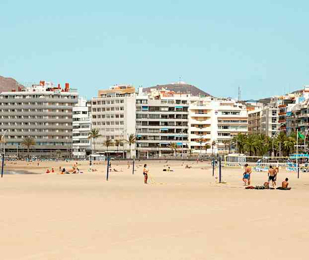Strand med sand og mennesker som soler seg og spiller volleyball foran flere høyhus. (AI-generert tekst)