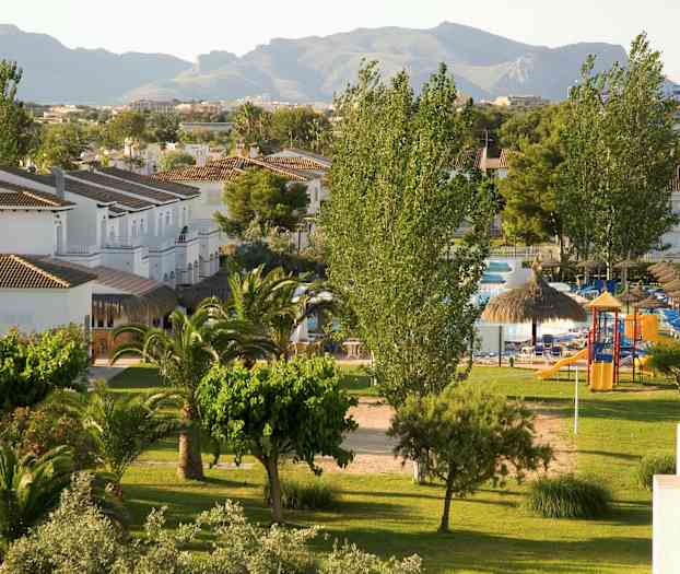 Utsikt over et grønt område med trær, plen og små hvite bygninger, samt bakgrunn med fjell. (AI-generert tekst)