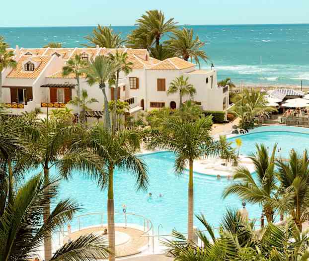 Hotellbygninger med terrakottafargede tak ved et basseng omgitt av palmer og hav i bakgrunnen. (AI-generert tekst)