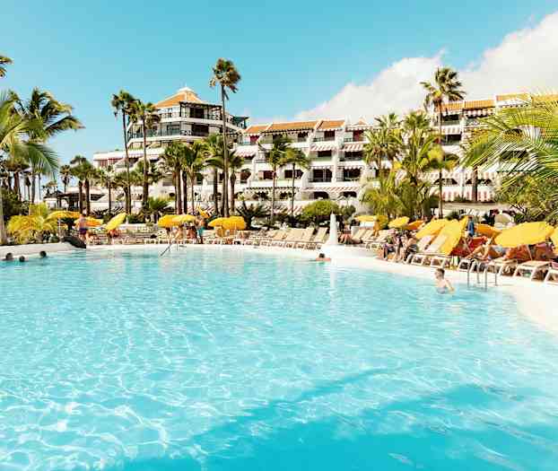 Stor basseng omgitt av solsenger og parasoller foran hotellbygning. (AI-generert tekst)