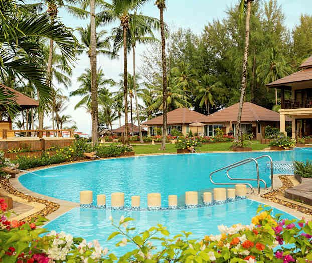 Ett ferieanlegg med et stort basseng i forgrunnen, omgitt av tropiske planter og palmer, med flere bungalower i bakgrunnen. (AI-generert tekst)
