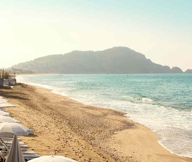 Strand med parasoller langs kysten og bakgrunn med fjell ved sjøen. (AI-generert tekst)