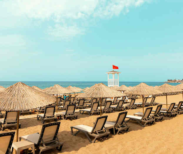 Strand med solsenger og parasoller samt et livreddertårn med rødt flagg. (AI-generert tekst)