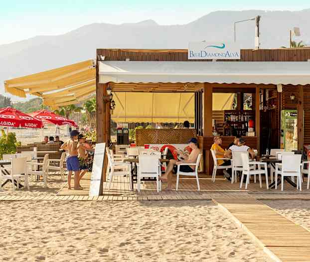 Strandrestaurant med uteservering og solsenger, fjell i bakgrunnen. (AI-generert tekst)