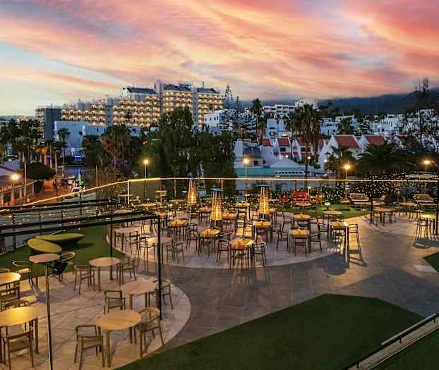 En utendørsrestaurant med bord og stoler under kveldsbelysning. I bakgrunnen vises bygninger og en fargerik solnedgang. (AI-generert tekst)
