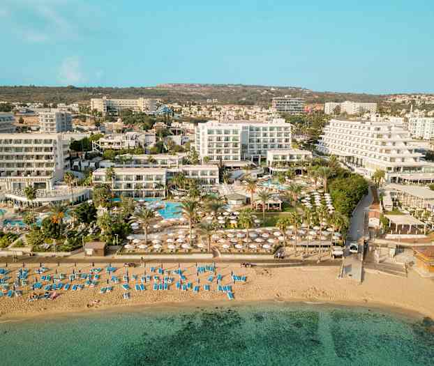 Luftfoto som viser en strand med solsenger og parasoller, samt hotellbygninger og bassengområde i bakgrunnen. (AI-generert tekst)