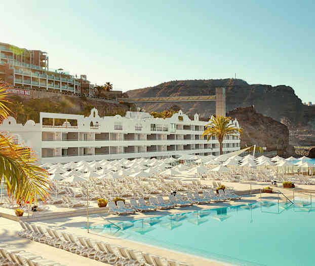 Hotellbygning med hvite fasader, solsenger og parasoller ved et stort basseng, samt klippeformasjoner i bakgrunnen. (AI-generert tekst)