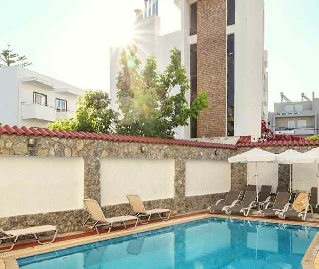 Et mindre utendørs svømmebasseng omgitt av solsenger og bygninger. (AI-generert tekst)