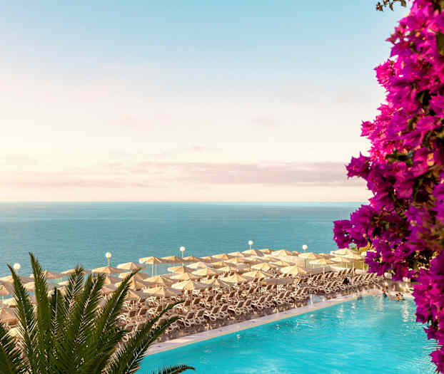 Solstoler med parasoller ved et basseng med utsikt over havet og blomster. (AI-generert tekst)